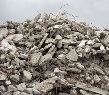 Ein großer Haufen aus grobem Bauschutt und Betonresten liegt unter freiem Himmel. Die Bruchstücke sind unterschiedlich groß und kantig, teils mit herausragenden Metallstäben und Armierungseisen. Der Himmel im Hintergrund ist grau und bewölkt, was die Szenerie nüchtern und industriell wirken lässt.

                          